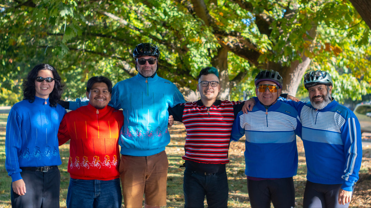 Six people standing together outdoors under a large leafy tree, wearing colorful cycling vintage sweaters and helmets. The group is posed in a sunny park setting.